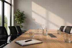 La imagen muestra una sala de reuniones moderna y luminosa. En el centro, hay una mesa de madera clara con un cuaderno y un bolígrafo, junto a varios vasos de agua y una jarra. Al fondo, se observa una planta verde y grandes ventanales que permiten la entrada de luz natural, creando un ambiente acogedor y profesional. La escena transmite una sensación de calma y preparación, ideal para discusiones importantes.